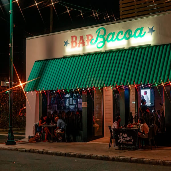 Bar Bacoa restaurant at night with a green awning, neon sign, and patrons dining outdoors.