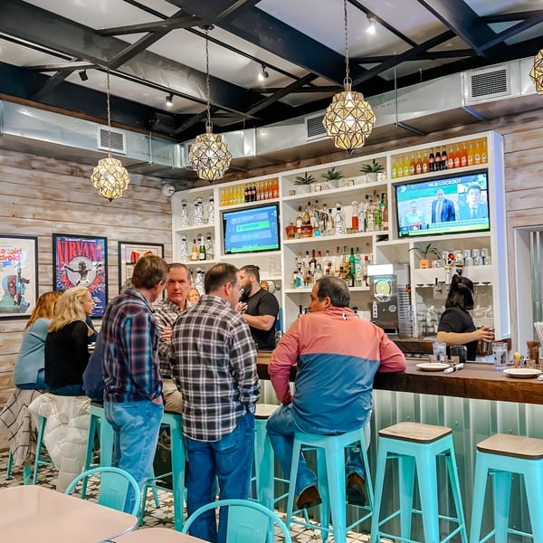 Patrons socialize at the Bar Bacoa bar, with a bartender serving drinks and TVs showing sports.