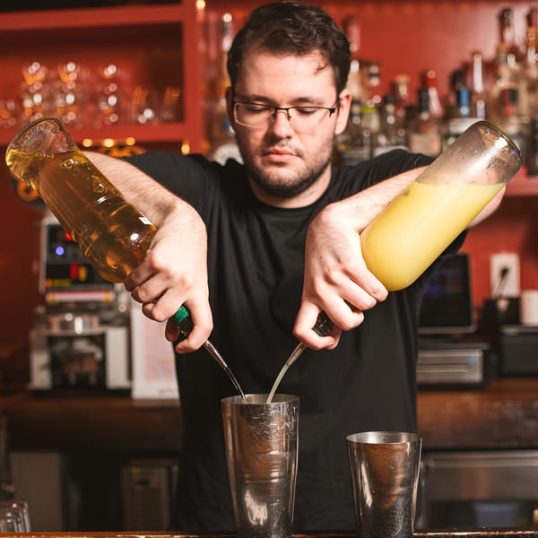 A Bar Bacoa bartender in glasses pours two liquids from bottles into a cocktail shaker.