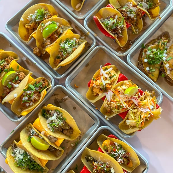 An overhead view of various Bar Bacoa tacos, including beef, cauliflower, and slaw, arranged on metal trays.