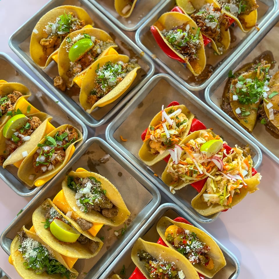 An overhead view of various Bar Bacoa tacos, including beef, cauliflower, and slaw, arranged on metal trays.