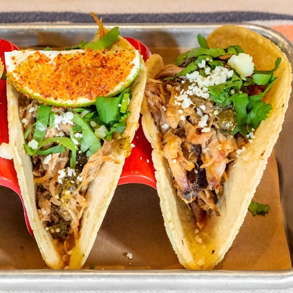 Two Bar Bacoa tacos with meat, cilantro, cheese, and a lime wedge on a red holder.