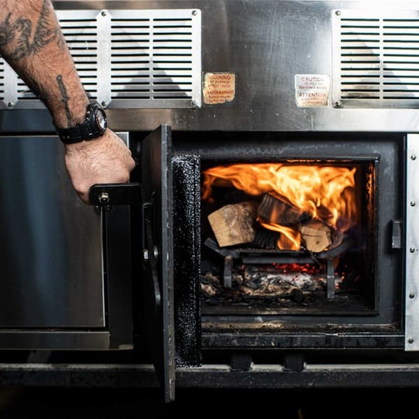 A chef's tattooed arm opening a wood-fired oven with burning logs inside, for Bar Bacoa's tacos.