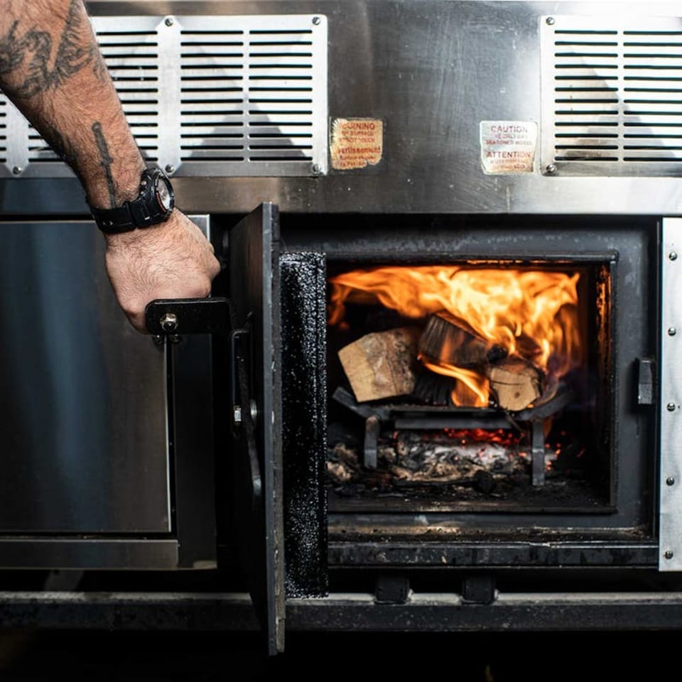 A chef's tattooed arm opening a wood-fired oven with burning logs inside, for Bar Bacoa's tacos.