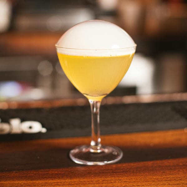 A vibrant yellow cocktail with a dome of white foam sits on a wooden bar top.