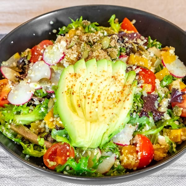 Colorful salad with sliced avocado, cherry tomatoes, radishes, quinoa, and pumpkin seeds in a black bowl.